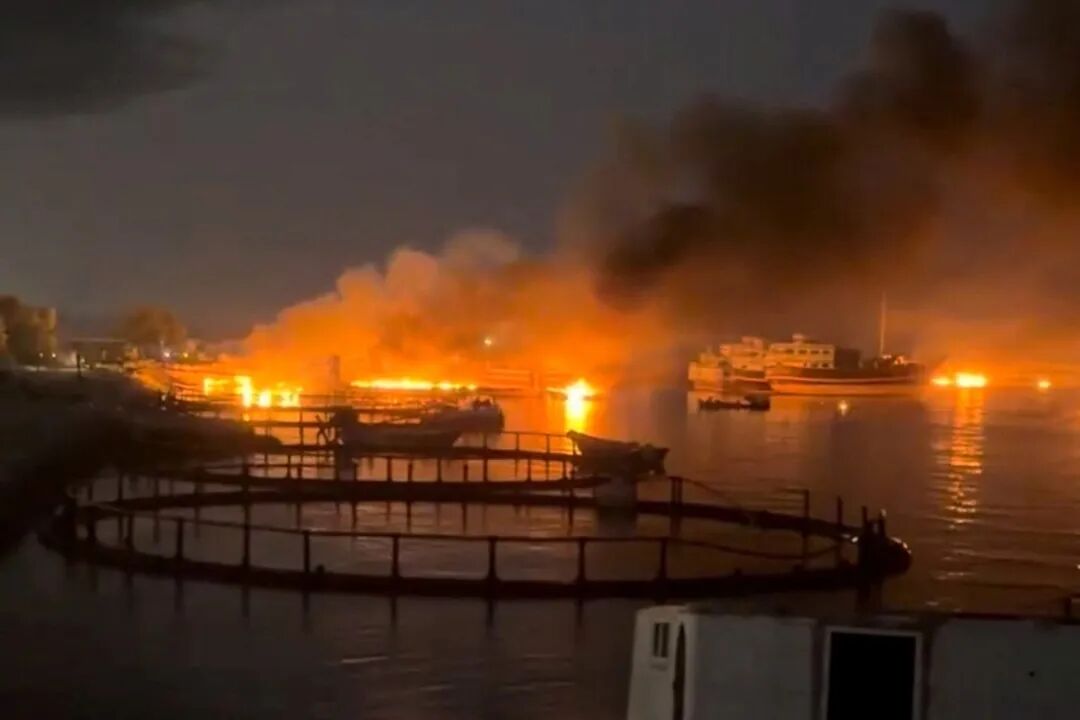 A nighttime view shows a large fire blazing along a shoreline, with intense orange flames and thick dark smoke rising into the sky. The fire reflects brightly on the water's surface. In the foreground, circular aquaculture pens float in the water, while several boats are positioned near the burning structures in the background.