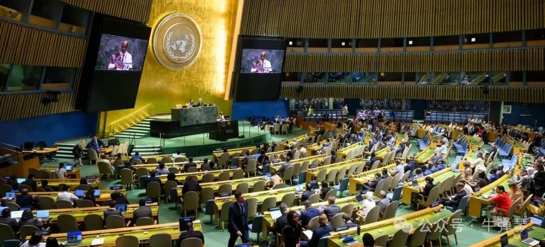 纽约United Nations全体会议厅内，代表们坐在弧形桌旁，面向中央讲台，讲台下方是一面金色的墙上的联合国徽章。